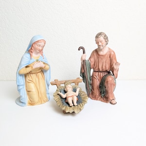 May include: A nativity set featuring three ceramic figurines. A kneeling figure in a light blue and yellow robe, a kneeling figure in a brown robe holding a staff, and a small infant figure lying in a brown manger with a blue interior lining.