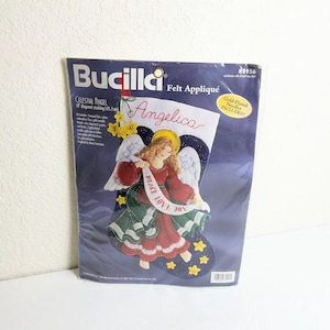 May include: A Bucilla Celestial Angel felt appliqué kit. The kit includes a 18-inch (45.7 cm) diagonal stocking design with an angel holding a banner that reads "Peace Love Joy". The kit includes gold-plated needles.