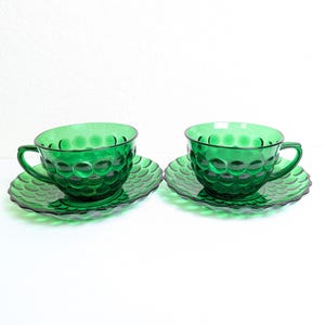 May include: Two emerald green glass teacups and saucers. The cups and saucers have a textured, bubble-like pattern. The cups have handles and sit on matching scalloped-edge saucers. The set is displayed on a white surface.