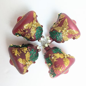 May include: Four burgundy-colored Christmas bell ornaments. Each bell is decorated with gold glitter, green glitter, and small red and gold accents. The bells are connected by a silver metal piece.