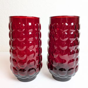 May include: Two ruby red drinking glasses with a textured, bubble-like pattern. The glasses have a cylindrical shape and a dark base. The vibrant red color and unique design make them a striking addition to any glassware collection.