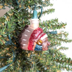 May include: A Native American-themed Christmas tree ornament. The ornament is a light bulb shaped like a Native American chief's head, with a red headdress, brown hair, and a detailed face. The ornament is hanging on a green Christmas tree.