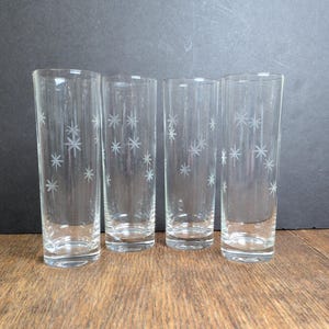 May include: Four clear glass tumblers with a starburst pattern etched on the surface. The glasses are tall and cylindrical, with a simple, elegant design. They are arranged in a row on a wooden surface.