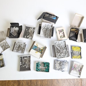 May include: An overhead shot displays several open boxes of sewing pins on a white surface. The pins are in various colors, including silver, yellow, and green. The boxes are open, revealing the pins inside.
