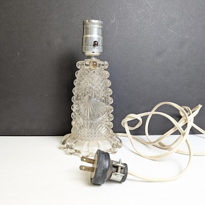 May include: A clear glass table lamp with a decorative, textured base and a silver-colored lamp holder. The lamp has a white cord and a black plug. The lamp is on a white surface with a dark gray background.