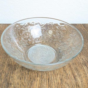 May include: A clear glass bowl with a floral pattern etched into the glass. The bowl has a wide, shallow shape and sits on a wooden surface. The floral design is repeated throughout the bowl's exterior and interior.