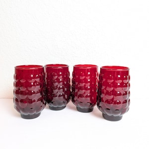 May include: Four vintage ruby red drinking glasses. Each glass has a textured, bubble-like pattern and a dark base. The glasses are cylindrical and appear to be made of glass.