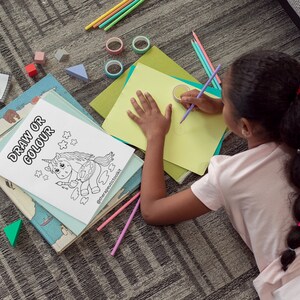 May include: A young person is coloring a black and white drawing of a unicorn holding a paintbrush and palette. The drawing is on a white sheet of paper with the words "Draw or Colour" at the top. The person is sitting on a gray carpet with a variety of colorful pencils and other art supplies scattered around them.