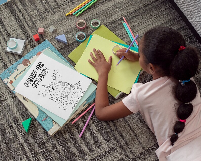 May include: A young person is coloring a black and white drawing of a unicorn holding a paintbrush and palette. The drawing is on a white sheet of paper with the words "Draw or Colour" at the top. The person is sitting on a gray carpet with a variety of colorful pencils and other art supplies scattered around them.