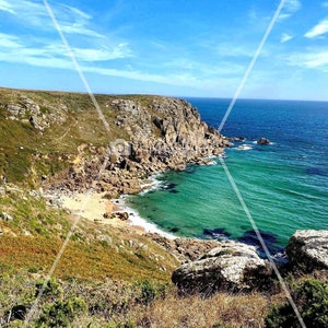 Pode incluir: Paisagem costeira c&eacute;nica com uma fal&eacute;sia rochosa que encontra o oceano turquesa sob um c&eacute;u azul brilhante. Uma pequena praia de areia est&aacute; aninhada numa enseada, com vegeta&ccedil;&atilde;o verde e rochas castanhas.