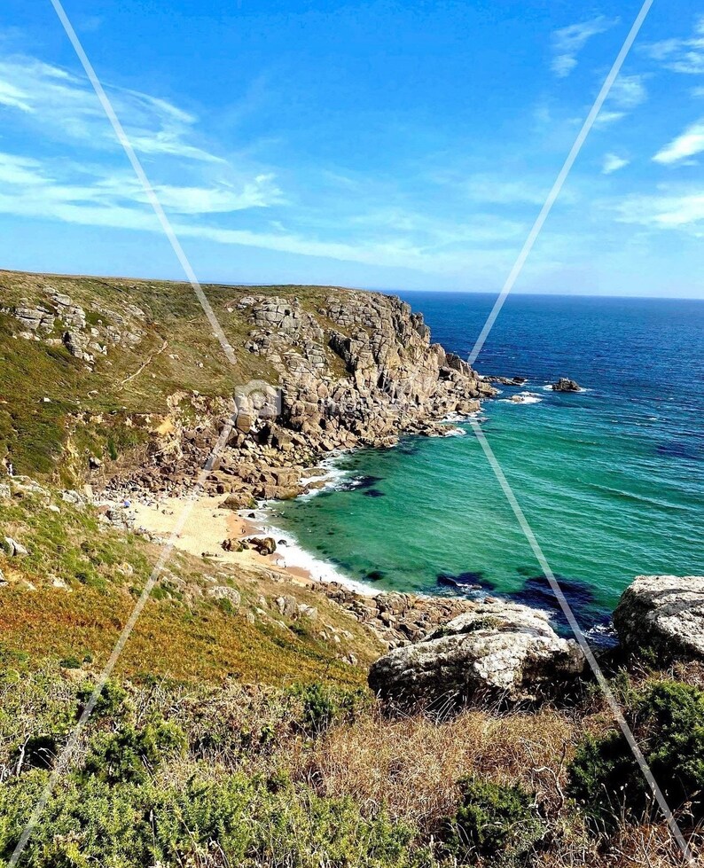 Pode incluir: Paisagem costeira c&eacute;nica com uma fal&eacute;sia rochosa que encontra o oceano turquesa sob um c&eacute;u azul brilhante. Uma pequena praia de areia est&aacute; aninhada numa enseada, com vegeta&ccedil;&atilde;o verde e rochas castanhas.