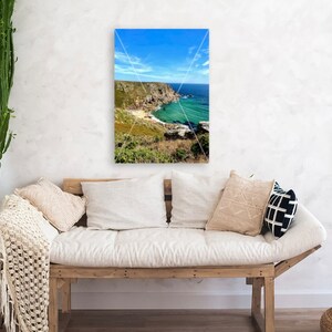 Pode incluir: Uma impress&atilde;o emoldurada de uma paisagem costeira com um c&eacute;u azul brilhante e &aacute;gua turquesa. A impress&atilde;o est&aacute; pendurada acima de um sof&aacute; de madeira com almofadas brancas e almofadas decorativas. Um cacto verde alto e uma cesta de vime tamb&eacute;m est&atilde;o na sala.