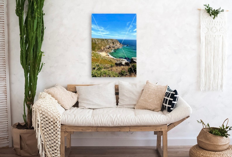 Pode incluir: Uma impress&atilde;o emoldurada de uma paisagem costeira com um c&eacute;u azul brilhante e &aacute;gua turquesa. A impress&atilde;o est&aacute; pendurada acima de um sof&aacute; de madeira com almofadas brancas e almofadas decorativas. Um cacto verde alto e uma cesta de vime tamb&eacute;m est&atilde;o na sala.