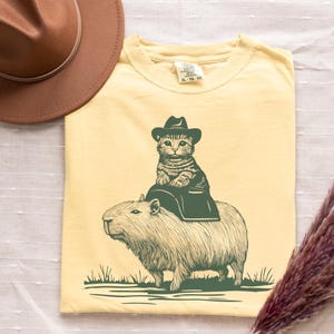 Può includere: T-shirt giallo chiaro con un'illustrazione stravagante di un gatto con un cappello da cowboy che cavalca un capibara. Il disegno è verde scuro su sfondo giallo chiaro. Un cappello da cowboy marrone è posto in alto a sinistra.