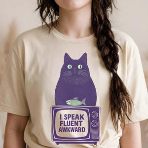 Può includere: Una t-shirt color crema con la grafica di un gatto viola scuro seduto sopra un televisore vintage. Lo schermo del televisore mostra il testo I SPEAK FLUENT AWKWARD e un piccolo scheletro di pesce verde è posizionato appena sopra.