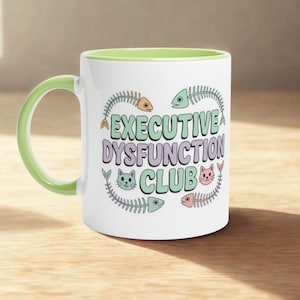 Peut inclure: Mug en céramique blanche avec une anse et un intérieur vert clair. Le mug présente le texte "EXECUTIVE DYSFUNCTION CLUB" dans une palette de couleurs pastel, ainsi que des illustrations de chats et d'arêtes de poisson. Le mug est sur une surface en bois.