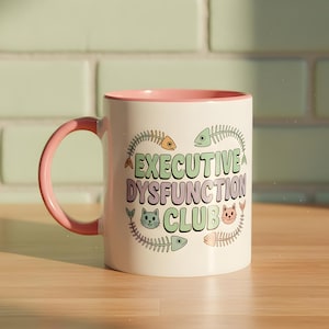 Peut inclure: Une tasse en céramique blanche avec un intérieur et une anse roses. La tasse porte l'inscription "EXECUTIVE DYSFUNCTION CLUB" dans une police fantaisiste, entourée d'arêtes de poisson et d'illustrations de chats. La tasse est posée sur une surface en bois.