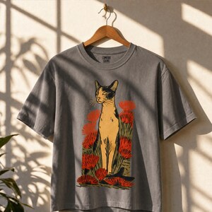 Pu&ograve; includere: Una t-shirt a maniche corte grigio antracite con una stampa grafica di un gatto beige e nero in piedi tra fiori rossi stilizzati e fogliame verde. L'etichetta interna sul collo riporta il testo Comfort Colors.