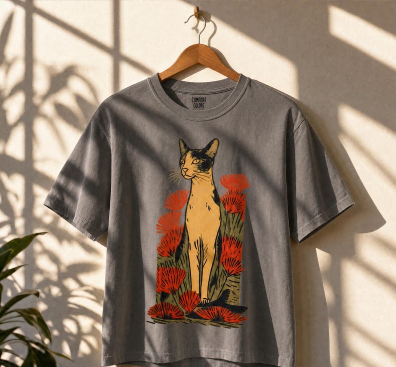 Pu&ograve; includere: Una t-shirt a maniche corte grigio antracite con una stampa grafica di un gatto beige e nero in piedi tra fiori rossi stilizzati e fogliame verde. L'etichetta interna sul collo riporta il testo Comfort Colors.