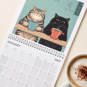 May include: A 2027 wall calendar for January featuring an illustration of a brown tabby cat holding a blue mug and a black cat holding an orange mug, both resting on a wooden counter against a teal background with a menu board and plant.
