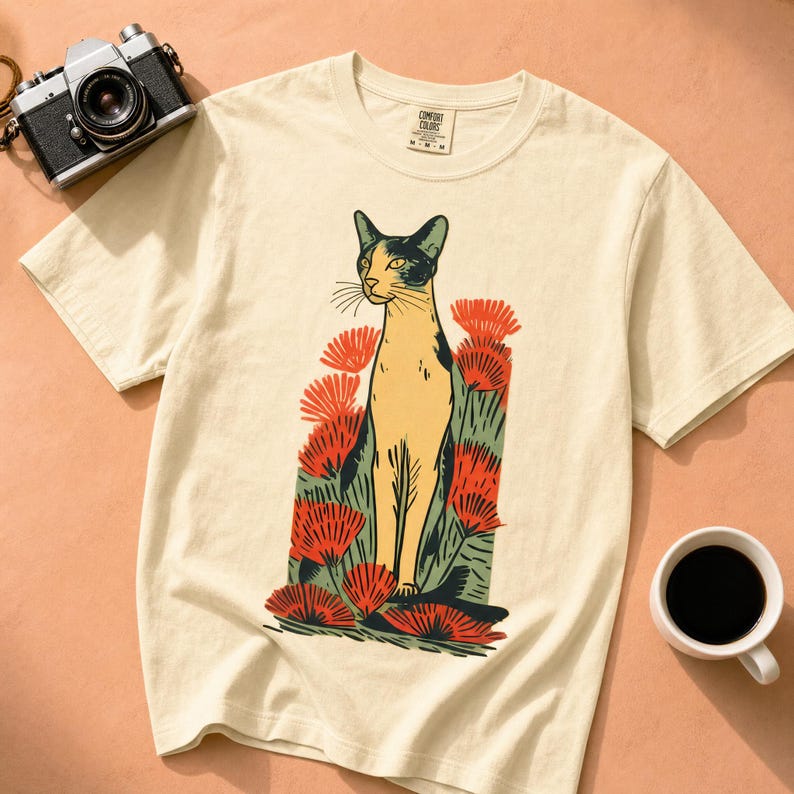 Pu&ograve; includere: Una t-shirt a maniche corte color crema con un'illustrazione grafica di un gatto in piedi tra fiori rossi e fogliame verde. La maglietta presenta un girocollo e un'etichetta del marchio Comfort Colors stampata all'interno del colletto.