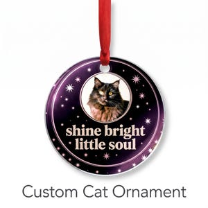 May include: A round, dark purple ornament featuring a central photograph of a fluffy black cat. The text "shine bright little soul" is printed in white below the cat. The background has a star pattern and a red ribbon for hanging.