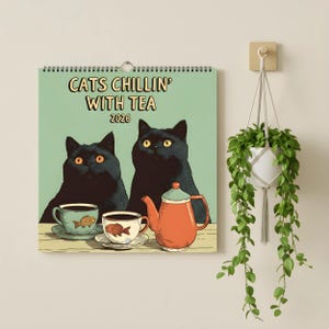 Può includere: Calendario da parete con l'illustrazione di due gatti neri, tazze da tè e una teiera. Il testo sul calendario recita "CATS CHILLIN' WITH TEA 2026". Il calendario è appeso a una parete accanto a una pianta sospesa.