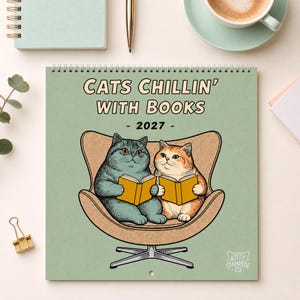 May include: A 2027 wall calendar titled CATS CHILLIN' WITH BOOKS featuring a light green cover with an illustration of a grey cat and an orange cat sitting in a tan chair, each holding an open yellow book. The logo KITTY STAMPEDE CO. is at the bottom.