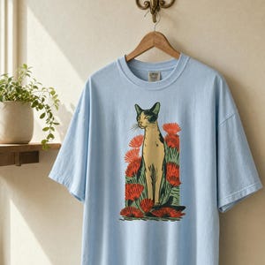 Pu&ograve; includere: Una t-shirt azzurro chiaro con una stampa grafica di un gatto in piedi tra fiori rossi. Il gatto &egrave; raffigurato in tonalit&agrave; crema e nero con accenti verdi. La maglietta presenta un girocollo e maniche corte.