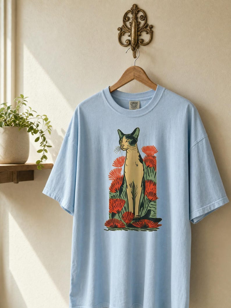 Pu&ograve; includere: Una t-shirt azzurro chiaro con una stampa grafica di un gatto in piedi tra fiori rossi. Il gatto &egrave; raffigurato in tonalit&agrave; crema e nero con accenti verdi. La maglietta presenta un girocollo e maniche corte.