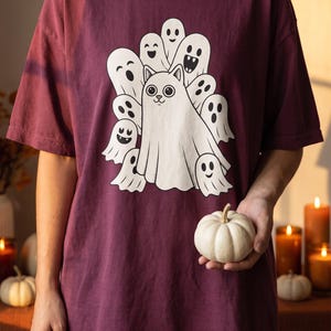 Può includere: Una t-shirt a maniche corte color bordeaux scuro con una stampa grafica centrale che raffigura un gatto bianco travestito da fantasma, circondato da diverse figure di fantasmi bianchi più piccoli con varie espressioni facciali.