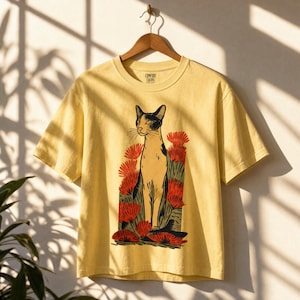 Pu&ograve; includere: Una t-shirt a maniche corte color giallo pallido con una stampa grafica di un gatto in piedi tra fiori rossi. La maglietta presenta un girocollo e un'etichetta del marchio Comfort Colors stampata all'interno del colletto.