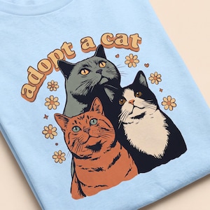 Può includere: Una t-shirt azzurro chiaro con una stampa grafica di tre gatti nei colori grigio, arancione e bianco e nero. Il testo "adopt a cat" è arcuato sopra i gatti in lettere arancioni, circondato da piccole illustrazioni di fiori gialli e puntini.