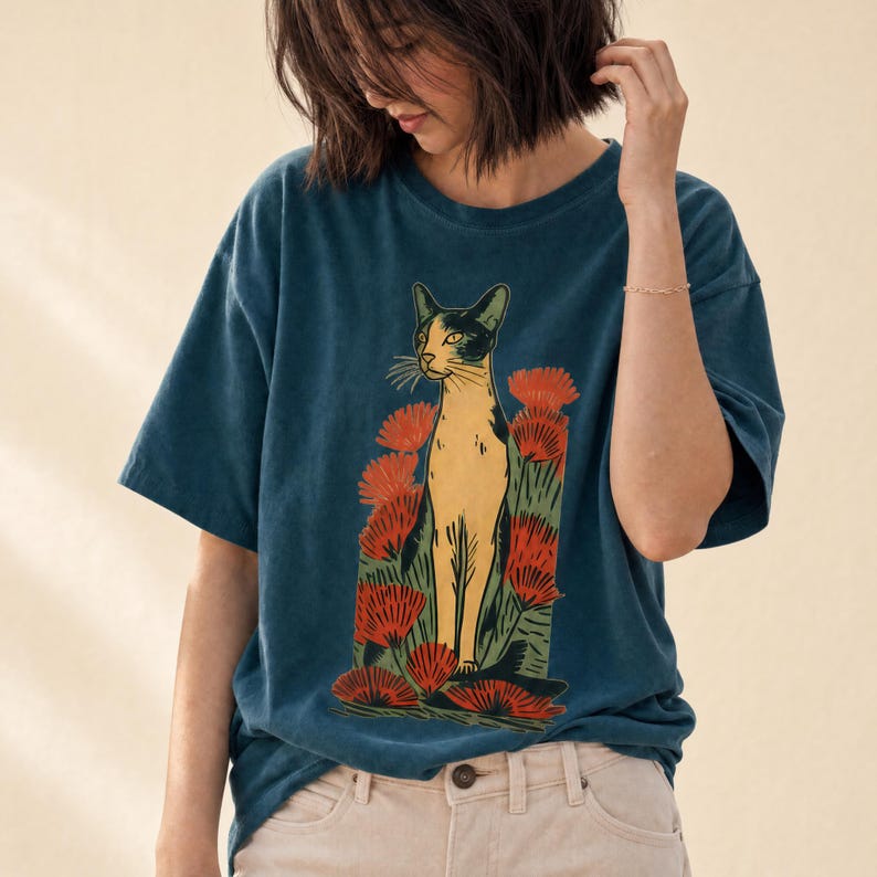Pu&ograve; includere: Una t-shirt a maniche corte blu scuro con una stampa grafica di un gatto beige e nero in piedi tra fiori rossi stilizzati e fogliame verde. Il gatto &egrave; raffigurato in posizione centrale con la testa leggermente girata di lato.