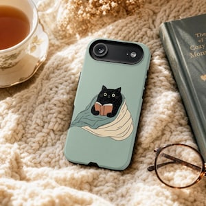May include: A sage green smartphone case featuring a graphic illustration of a black cat wrapped in a blue and cream-colored blanket while reading an open book. The case has a black camera cutout at the top and a smooth, matte-finish surface.