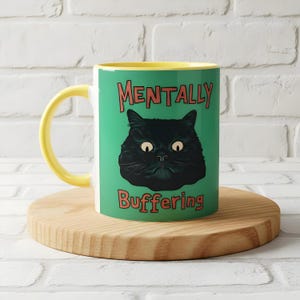May include: A ceramic coffee mug with a yellow handle and interior. The mug features a black cat illustration on a teal background, with the words "MENTALLY" and "BUFFERING" in red text.