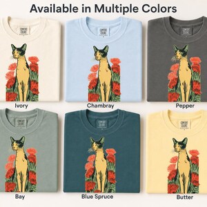 Pu&ograve; includere: Sei magliette nei colori avorio, chambray, pepe, baia, abete blu e burro. Ogni maglietta presenta una grafica di un gatto in piedi tra fiori rossi. Il testo "Available in Multiple Colors" appare in alto, con i nomi dei colori sotto.