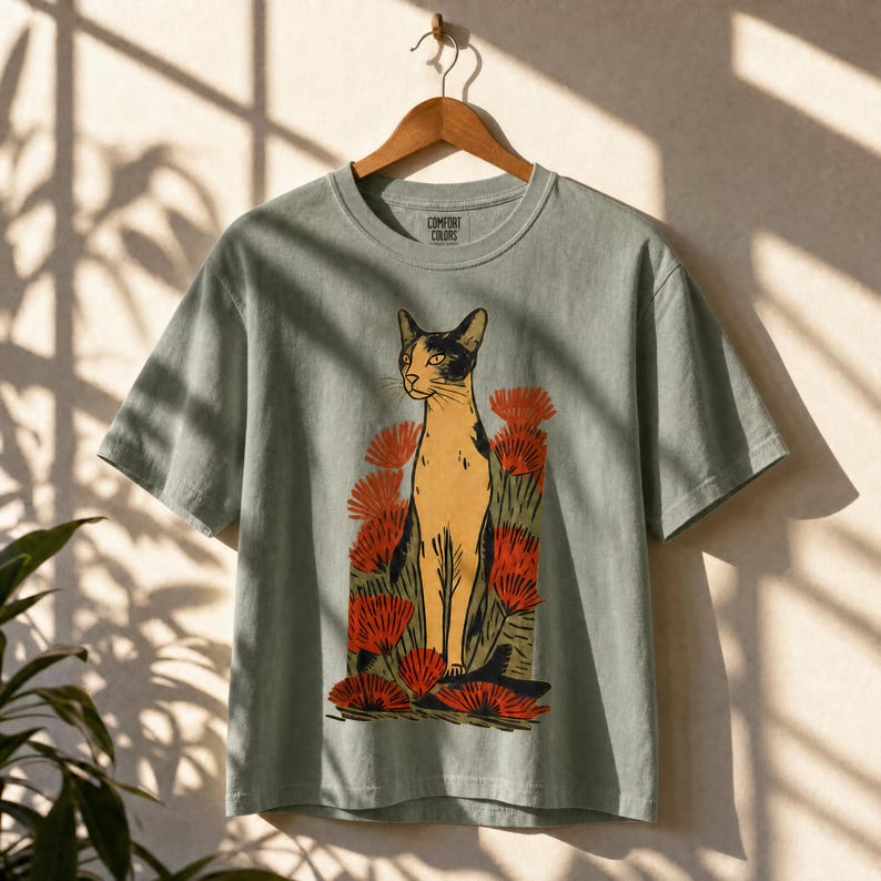 Pu&ograve; includere: Una t-shirt a maniche corte color verde salvia con una stampa grafica di un gatto beige e nero in piedi tra fiori rossi stilizzati. L'etichetta interna sul collo riporta il testo Comfort Colors. La maglietta &egrave; appesa a una gruccia.