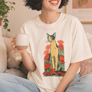 Pu&ograve; includere: Una t-shirt a maniche corte color crema con una stampa grafica di un gatto in piedi tra un gruppo di fiori rossi vivaci e fogliame verde. Il gatto &egrave; raffigurato in tonalit&agrave; gialle e nere con un'estetica stilizzata da stampa a blocchi.