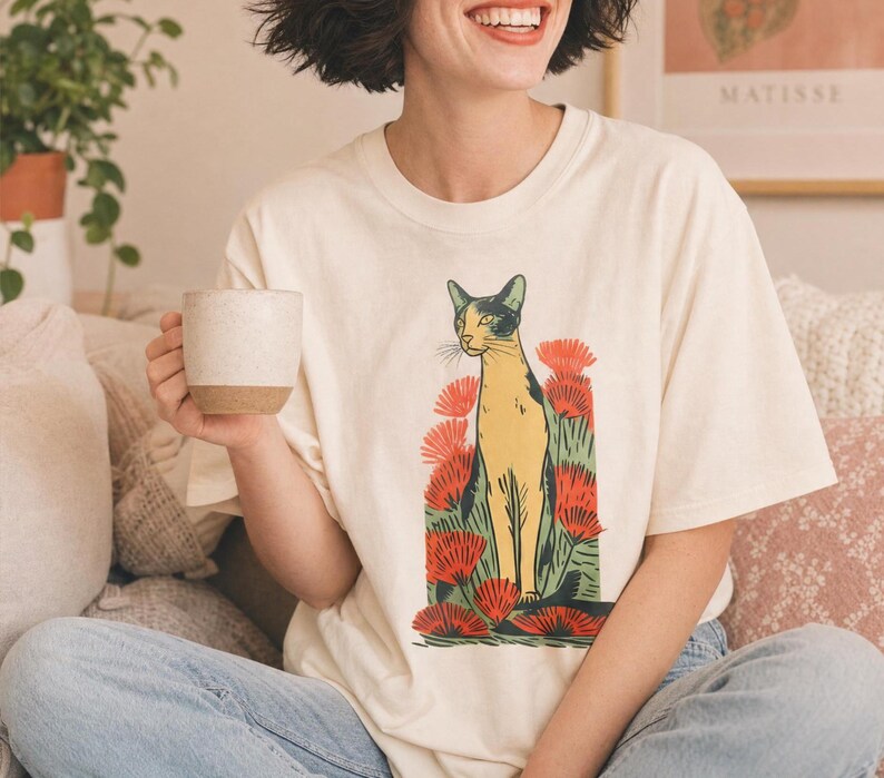 Pu&ograve; includere: Una t-shirt a maniche corte color crema con una stampa grafica di un gatto in piedi tra un gruppo di fiori rossi vivaci e fogliame verde. Il gatto &egrave; raffigurato in tonalit&agrave; gialle e nere con un'estetica stilizzata da stampa a blocchi.