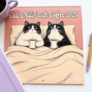 May include: A 2027 wall calendar titled Cats Chillin' with Coffee featuring an illustration of two black and white tuxedo cats sitting up in bed under a beige blanket, each holding a white coffee mug. The background is a soft peach color.