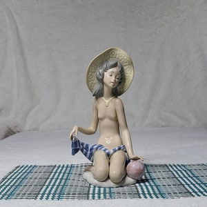 Puede incluir: Una figurita de porcelana de una niña que lleva un sombrero de paja y una bufanda a cuadros azules y blancos, sentada en una manta a cuadros con una pelota rosa.