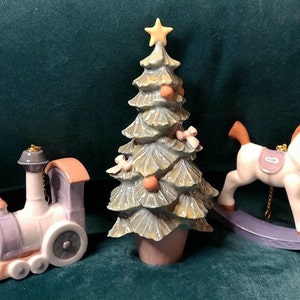 May include: Three porcelain Christmas ornaments: a blue and pink train, a rocking horse with a pink saddle, and a green and brown Christmas tree with a yellow star on top.