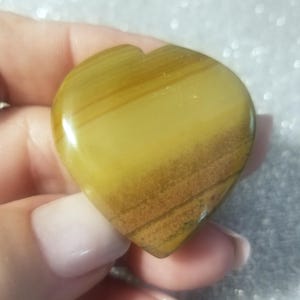 May include: A heart-shaped stone with a polished surface. The stone features bands of yellow and brown hues, creating a natural, earthy appearance. The heart shape is smooth and symmetrical, perfect for a pendant or decorative item.