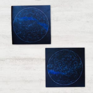 May include: Two square black wall art pieces featuring circular star charts. Each print displays white constellations and celestial lines within a circle, accented by a horizontal band of deep blue color across the center of the star map design.