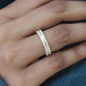 May include: A gold ring featuring a zigzag design of rectangular, clear gemstones. The ring is worn on a finger, with the skin tone visible in the background. The ring's design is a continuous band of alternating gemstones.