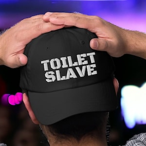 May include: A black baseball cap with a white embroidered design that says "TOILET SLAVE".