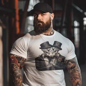 May include: A white t-shirt featuring a graphic of two skeletons in tuxedos, one holding a rose. The skeletons are wearing top hats and bow ties. The man is wearing a black cap and has a beard.