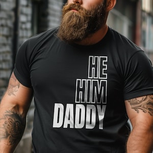 May include: Black t-shirt with white text that reads "HE HIM DADDY".