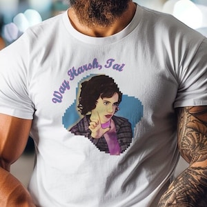 Peut inclure: T-shirt blanc avec une image pixélisée d'une femme aux cheveux foncés portant une chemise violette. Le texte "Way Harsh, Tai" est imprimé en violet au-dessus de l'image.